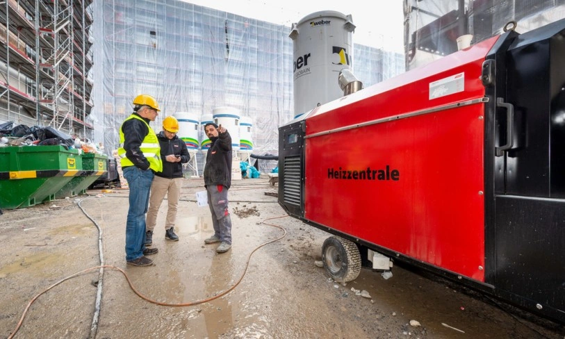 Rotes Dieselheizgerät auf einer Baustelle. Mit Bauarbeitern welche daneben stehen.